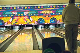 A bowler at the foul line on Flamingo Bowl's lanes, with colorful lane wall art and a full rack of pins.